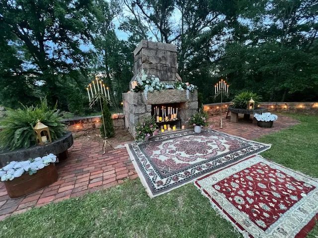 Outdoor memorial scene with a stone fireplace, candles, and floral garlands, set on brick patio with Persian-style rugs and greenery nearby.