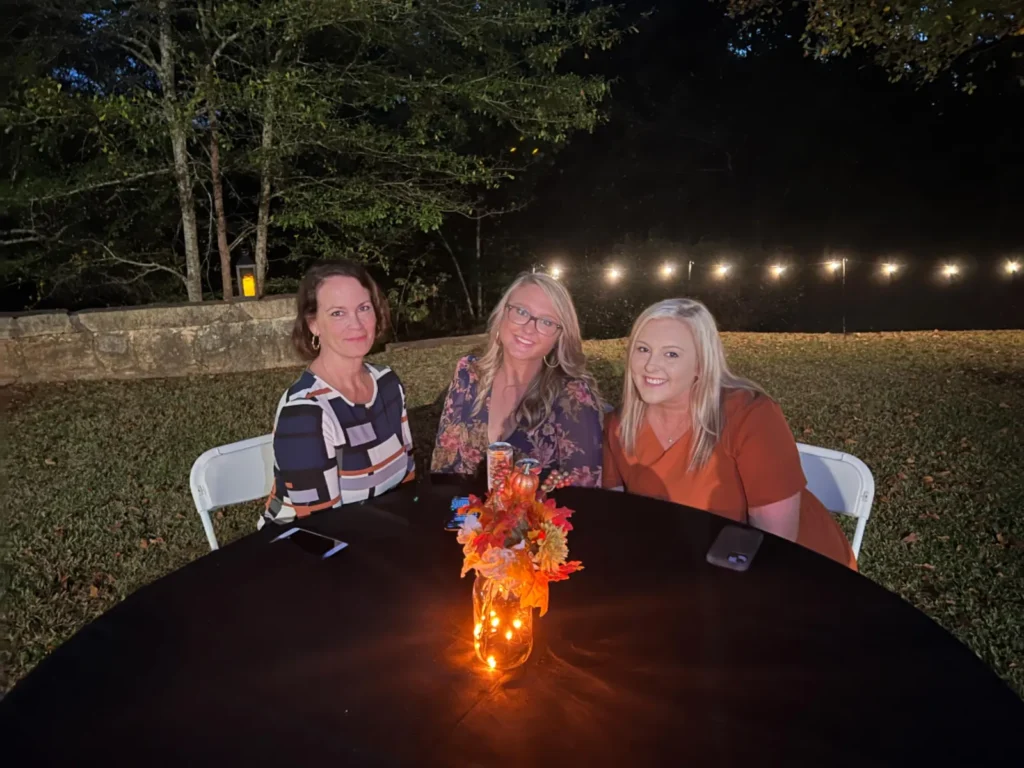 three women sit at a dark outdoor table with a lit jar centerpiece and autumn leaves, smiling at the camera.