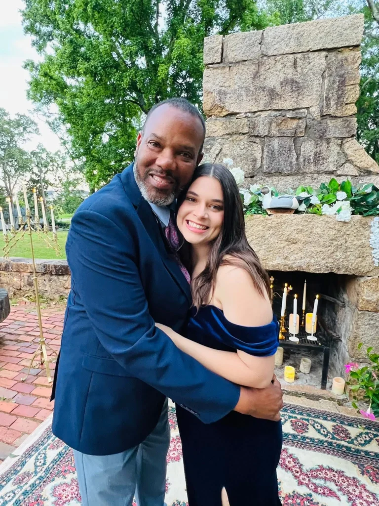 A couple embraces and smiles at an outdoor setting with a stone fireplace behind them.