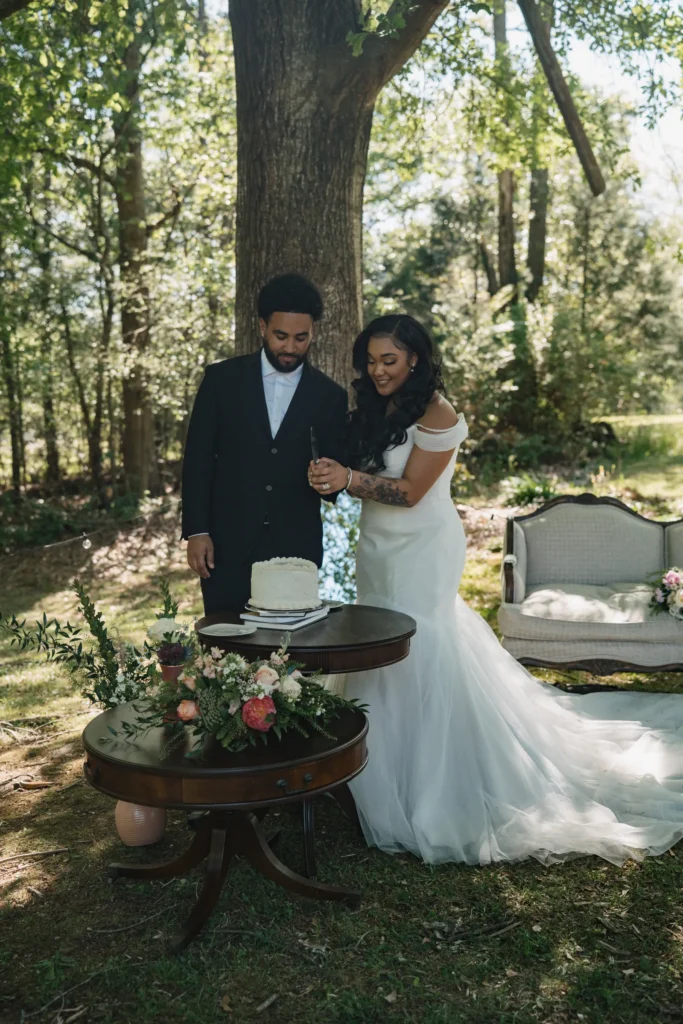 outdoor-cake-cutting-ceremony - Riegel Hall - Ware Shoals, SC bride and groom in formal attire cut a white wedding cake outdoors under a large tree, with floral decor nearby.
