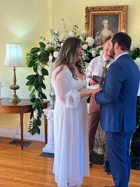 wedding-ceremony-indoor-venue - Riegel Hall - Ware Shoals, SC bride in a white dress and groom in a blue suit exchange vows during an indoor ceremony, with an officiant and a lush white floral arrangement behind them.